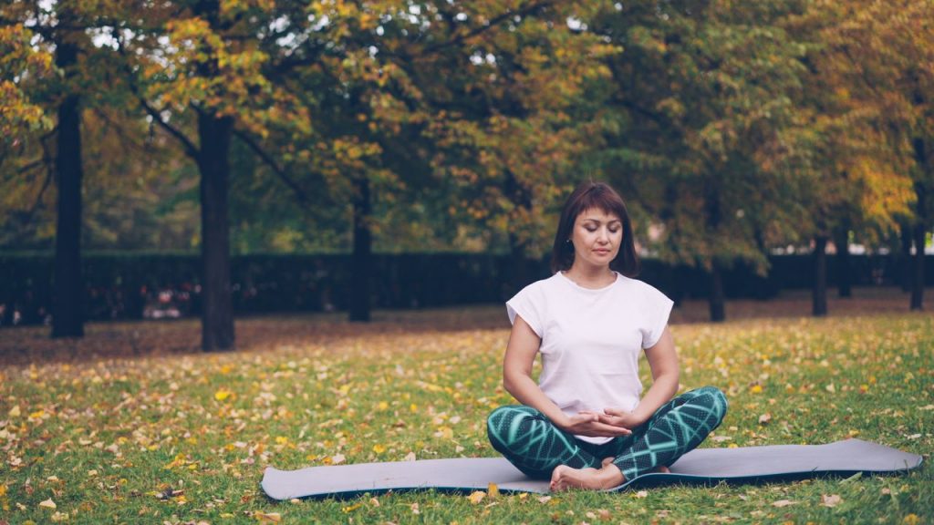 A woman practicing meditation on a yoga mat in a serene autumn park setting.
