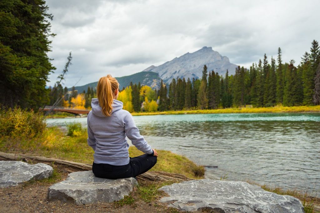 Woman meditating by a serene river with mountains in the background during autumn.