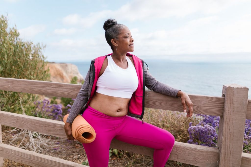 Confident senior woman in activewear holding a yoga mat, enjoying a scenic outdoor view by the ocean.