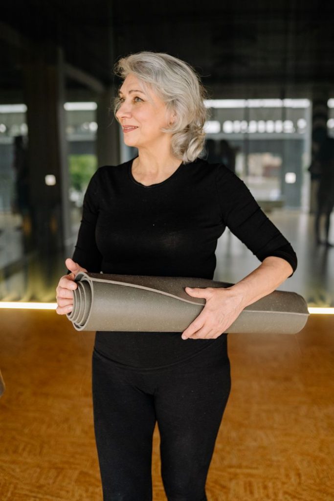 Elderly woman in activewear smiling and holding a yoga mat indoors.
